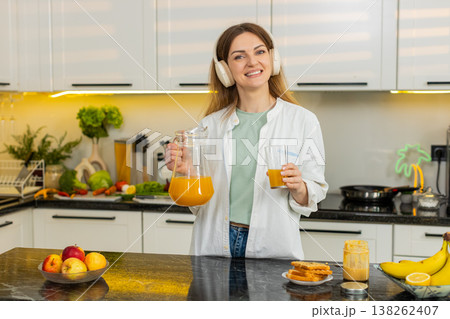 Middle-aged woman dancing with headphones in kitchen drinking fresh orange juice for diet wellness Middle-aged woman dancing with headphones in kitchen drinking fresh orange juice for diet wellness 138262407