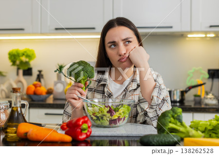 Exhausted young woman struggles with keto diet while eating green salad at kitchen table quietly now 138262528