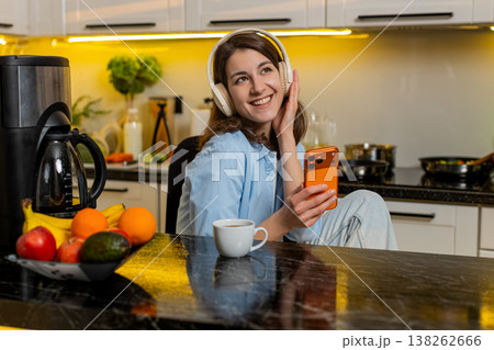 Young woman relaxing in home kitchen with headphones enjoying music while checking smartphone screen Young woman relaxing in home kitchen with headphones enjoying music while checking smartphone screen 138262666
