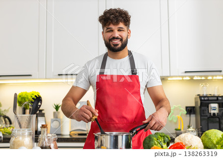 Arabian young adult man smiling stirring soup pot on stove in home kitchen for healthy dinner recipe 138263319