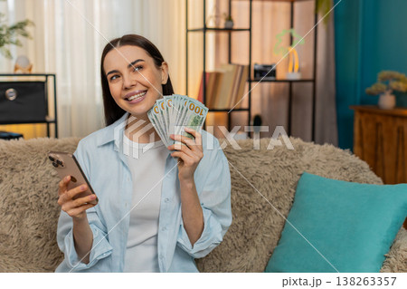 Young woman on sofa holding cash dollars and smartphone thoughtfully planning spending choices Young woman on sofa holding cash dollars and smartphone thoughtfully planning spending choices 138263357