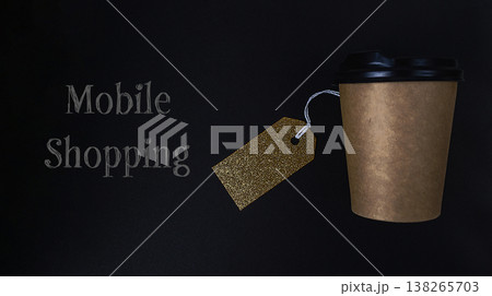 Brown paper coffee cup with black lid and golden tag on dark background, representing mobile shopping and discounts for modern consumers seeking convenience and savings Brown paper coffee cup with black lid and golden tag on dark background, representing mobile shopping and discounts for modern consumers seeking convenience and savings 138265703