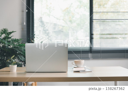 Minimal coworking desk with laptop, coffee cup and notebook beside small plant by window blinds, clean modern office workspace with copyspace for text and branding, productivity, remote work concept. 138267362