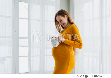 Pregnancy motherhood people expectation future. Pregnant woman with big belly holding newborn baby booties smiling at home. Young mom enjoying pregnancy. Maternity tenderness parenthood new life 138267399