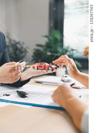 Agent presenting red model car and pen to client above contract with car key on office desk, representing vehicle purchase consultation, ownership planning, dealership service and trust. Agent presenting red model car and pen to client above contract with car key on office desk, representing vehicle purchase consultation, ownership planning, dealership service and trust. 138267503