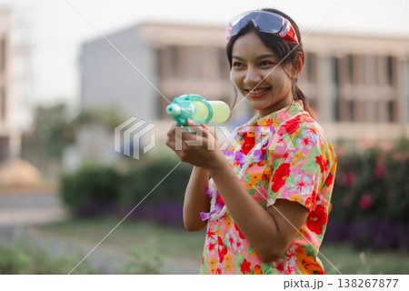 Smiling young woman in floral shirt holding water gun during Songkran celebration outdoors, wearing splash goggles with white powder on her face, capturing playful summer holiday energy. Smiling young woman in floral shirt holding water gun during Songkran celebration outdoors, wearing splash goggles with white powder on her face, capturing playful summer holiday energy. 138267877