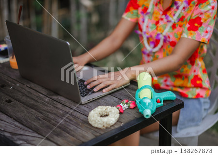 Detail shot of hands typing on laptop with water gun and flower garland on table, illustrating Songkran workspace, seasonal remote work, digital routine and festive summer lifestyle. Detail shot of hands typing on laptop with water gun and flower garland on table, illustrating Songkran workspace, seasonal remote work, digital routine and festive summer lifestyle. 138267878