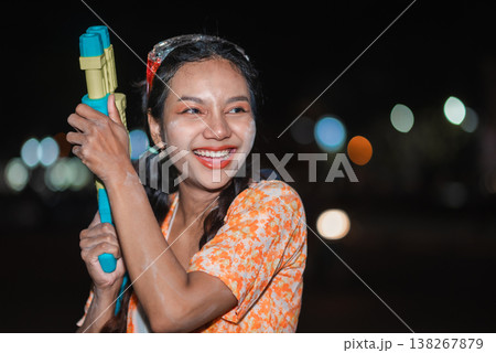 Portrait of smiling young woman holding water gun during Songkran night celebration, with wet face, soft bokeh lights and festive styling, showing cheerful Thai holiday mood, street fun and summer Portrait of smiling young woman holding water gun during Songkran night celebration, with wet face, soft bokeh lights and festive styling, showing cheerful Thai holiday mood, street fun and summer 138267879