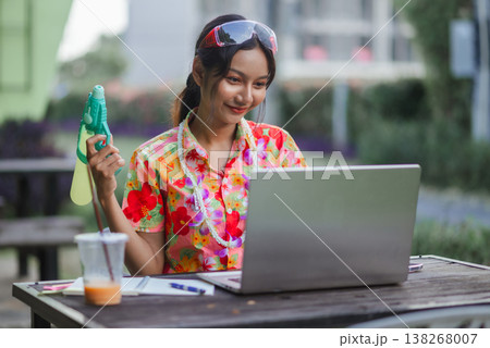 Young woman holding water gun while using laptop outdoors during Songkran festival, representing remote work, festive mood, summer lifestyle, digital freedom and modern holiday productivity. Young woman holding water gun while using laptop outdoors during Songkran festival, representing remote work, festive mood, summer lifestyle, digital freedom and modern holiday productivity. 138268007