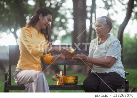 Young woman and elderly woman sharing a traditional Songkran water blessing on a bench outdoors, capturing Thai family values, respect for elders and a calm moment of cultural connection. Young woman and elderly woman sharing a traditional Songkran water blessing on a bench outdoors, capturing Thai family values, respect for elders and a calm moment of cultural connection. 138268070