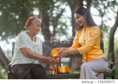 Young woman pouring water into an elderly woman hands on a wooden bench during Songkran, showing Thai family values, respect for elders and a peaceful traditional New Year blessing ritual. Young woman pouring water into an elderly woman hands on a wooden bench during Songkran, showing Thai family values, respect for elders and a peaceful traditional New Year blessing ritual. 138268071
