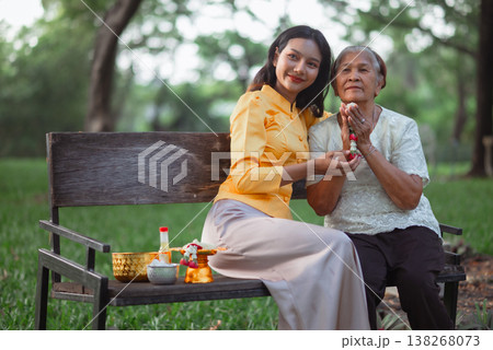 Young woman and elderly woman sitting together on a bench with a jasmine garland during Songkran, portraying Thai family bonding, respect for elders and warm cultural tradition with copy space. Young woman and elderly woman sitting together on a bench with a jasmine garland during Songkran, portraying Thai family bonding, respect for elders and warm cultural tradition with copy space. 138268073