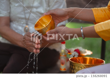 Close shot of a young woman gently pouring water from a golden bowl onto an elderly woman hands during a Thai traditional blessing ceremony, representing care, respect and cultural heritage. Close shot of a young woman gently pouring water from a golden bowl onto an elderly woman hands during a Thai traditional blessing ceremony, representing care, respect and cultural heritage. 138268091