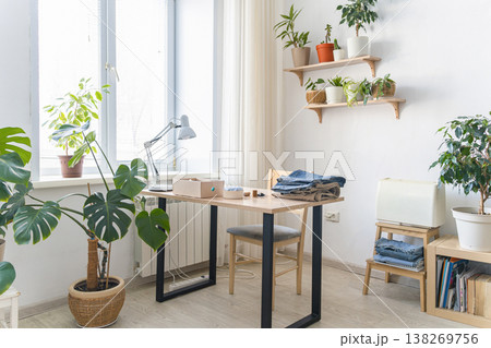 Cozy home workspace with houseplants and hobby items. Bright home office featuring a desk with sewing supplies, folded fabric, a sewing machine, and lush green houseplants Cozy home workspace with houseplants and hobby items. Bright home office featuring a desk with sewing supplies, folded fabric, a sewing machine, and lush green houseplants 138269756