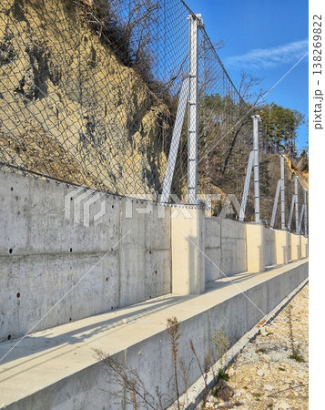Concrete retaining wall with steel mesh fence for landslide and rockfall protection on a steep slope. 138269822