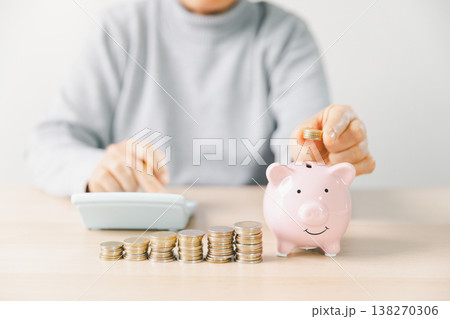 Hand placing a coin into a pink piggy bank beside stacks of money while writing budget notes at home. Personal finance, savings plan, budgeting, investment, long-term future security goals. Hand placing a coin into a pink piggy bank beside stacks of money while writing budget notes at home. Personal finance, savings plan, budgeting, investment, long-term future security goals. 138270306