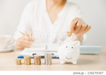 Hand placing a coin into a pink piggy bank beside stacks of money while writing budget notes at home. Personal finance, savings plan, budgeting, investment, long-term future security goals. Hand placing a coin into a pink piggy bank beside stacks of money while writing budget notes at home. Personal finance, savings plan, budgeting, investment, long-term future security goals. 138270307
