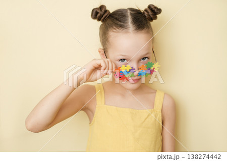 The cute girl lowered glasses decorated with pieces of colorful puzzles, looking suspiciously and smiling at the camera. World Autism Awareness Day. Education and social equality The cute girl lowered glasses decorated with pieces of colorful puzzles, looking suspiciously and smiling at the camera. World Autism Awareness Day. Education and social equality 138274442