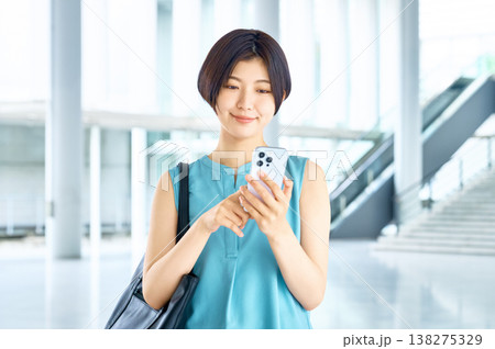 オフィスビルや駅の構内でスマートフォンを操作しながら微笑む若い女性 オフィスビルや駅の構内でスマートフォンを操作しながら微笑む若い女性 138275329