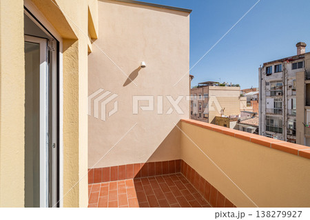 A sunny balcony with a terracotta floor and beige walls. The view includes nearby buildings under a clear blue sky. A sunny balcony with a terracotta floor and beige walls. The view includes nearby buildings under a clear blue sky. 138279927