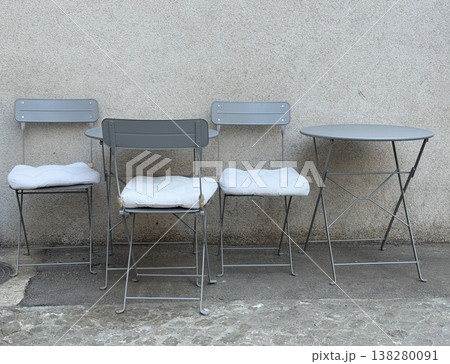 Gray folding chairs and tables arranged against textured wall in outdoor cafe area. Minimal hospitality setup, urban seating design, small business terrace and informal social gathering space 138280091