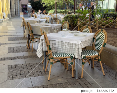 Restaurant terrace with set tables and woven chairs along a colonnade in city light. European outdoor dining, hospitality service, reservation setup and elegant sidewalk seating for guests Restaurant terrace with set tables and woven chairs along a colonnade in city light. European outdoor dining, hospitality service, reservation setup and elegant sidewalk seating for guests 138280112