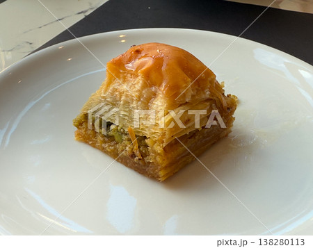 Pistachio baklava portion served on a white plate with crisp layered phyllo and glossy syrup. Traditional dessert plating, sweet hospitality, menu illustration and closeup food presentation Pistachio baklava portion served on a white plate with crisp layered phyllo and glossy syrup. Traditional dessert plating, sweet hospitality, menu illustration and closeup food presentation 138280113