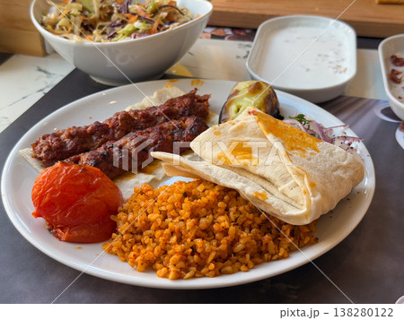 Adana kebab plate with bulgur pilaf, lavash, roasted tomato and grilled pepper. Traditional Turkish lunch serving, spicy minced meat entree and restaurant menu presentation with sides Adana kebab plate with bulgur pilaf, lavash, roasted tomato and grilled pepper. Traditional Turkish lunch serving, spicy minced meat entree and restaurant menu presentation with sides 138280122