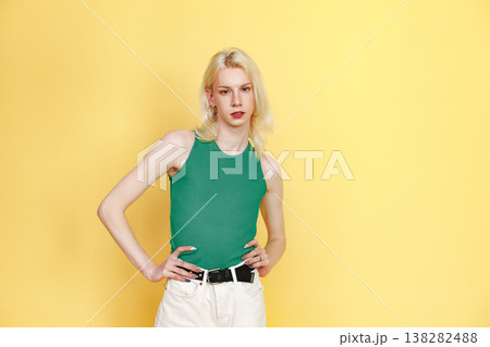 Young blonde man with make up on his face posing for a photo. Man wearing fashionable green top and has red lips. Man isolated on yellow background. 138282488