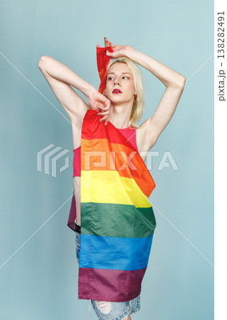 Young blonde man with make up on his face and rainbow flag posing for a photo. Man wearing fashionable pink t-shirt and has red lips. Man isolated on blue background. Young blonde man with make up on his face and rainbow flag posing for a photo. Man wearing fashionable pink t-shirt and has red lips. Man isolated on blue background. 138282491