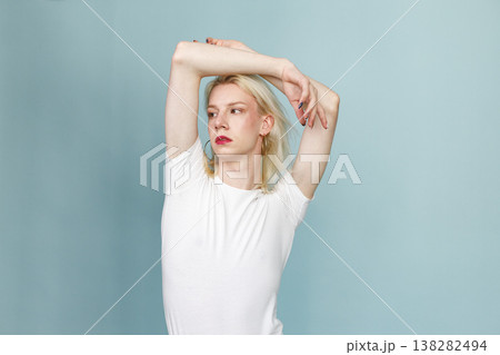 Young blonde man with make up on his face posing for a photo. Man wearing fashionable white t-shirt and has red lips. Man isolated on blue background. 138282494