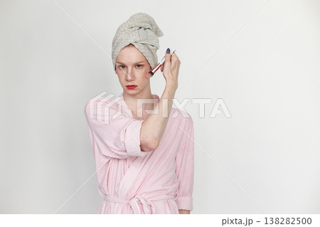 Young man with make up on his face holding brush for make up. Man wearing pink dressing gown and has a towel on his head. Man isolated on white background. Young man with make up on his face holding brush for make up. Man wearing pink dressing gown and has a towel on his head. Man isolated on white background. 138282500