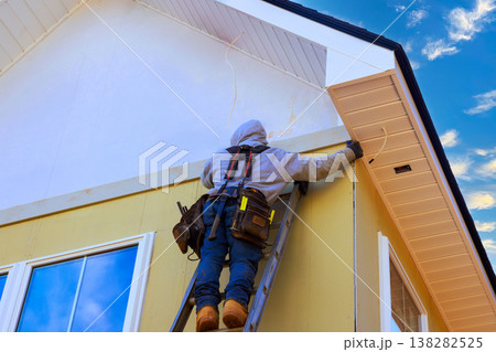 Worker is using ladder to fix trim on house while installing fiber siding panels Worker is using ladder to fix trim on house while installing fiber siding panels 138282525