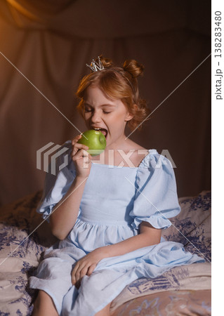 Little girl in blue dress with crown eating green apple, princess concept, studio drapery background 138283480