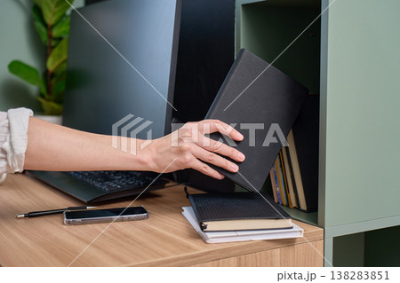 Hand placing black notebook on shelf beside desk with monitor, smartphone and journals 138283851
