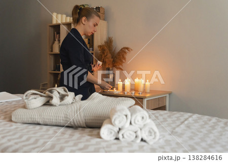 Massage therapist placing lit candles in spa treatment room, preparing for relaxing wellness session with towels robe on massage bed. Focus on comfort Massage therapist placing lit candles in spa treatment room, preparing for relaxing wellness session with towels robe on massage bed. Focus on comfort 138284616