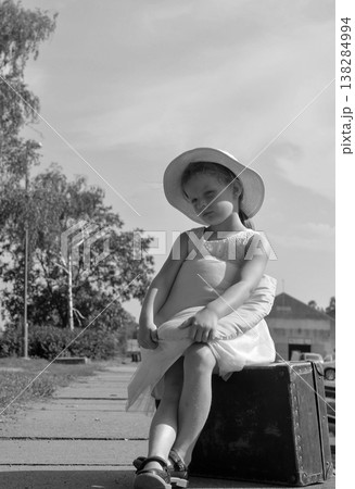 Young girl in a hat sits on an old suitcase, waiting patiently outdoors 138284994