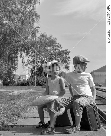 Two children in vintage clothing wait at a train station with luggage Two children in vintage clothing wait at a train station with luggage 138284996