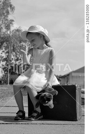 Young girl in a hat and dress sits on a suitcase with a stuffed toy, waiting for a journey 138285000