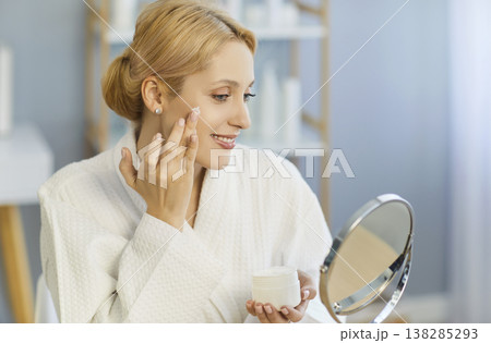 Smiling young woman in bathrobe applying face cream and looking in mirror during skincare routine. Smiling young woman in bathrobe applying face cream and looking in mirror during skincare routine. 138285293