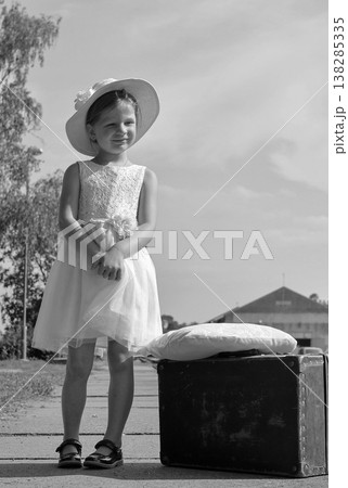 Young girl in a white dress and hat standing next to a vintage suitcase on a sunny day 138285335