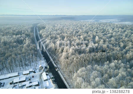Aerial view of winter landscape with snow covered woods and black asphalt forest road on cold wintry day Aerial view of winter landscape with snow covered woods and black asphalt forest road on cold wintry day 138288278