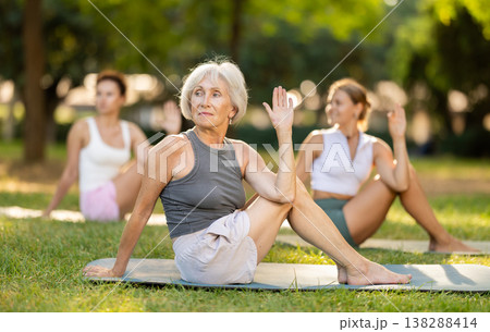 Sedulous old lady and group of women doing spinal twist pose of yoga on mat in light city park together Sedulous old lady and group of women doing spinal twist pose of yoga on mat in light city park together 138288414