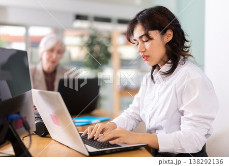 Young woman sitting at desk and typing on laptop in modern office Young woman sitting at desk and typing on laptop in modern office 138292136
