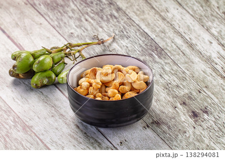 Roasted cashew nuts in a brown bowl with a cluster of raw green cashew fruit on a rustic wooden table. 138294081