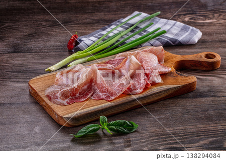 Thinly sliced cured meat, possibly prosciutto, on a wooden cutting board with green onions and basil. Thinly sliced cured meat, possibly prosciutto, on a wooden cutting board with green onions and basil. 138294084