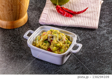 Homemade guacamole dip in a ceramic bowl with fresh avocado, chili peppers, and a wooden mortar on a dark background. 138294109
