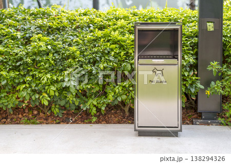 A modern stainless steel pet waste bin with a dog icon stands on a concrete path in front of a lush green hedge. It features a disposal drawer and a nearby poop bag dispenser for public use. A modern stainless steel pet waste bin with a dog icon stands on a concrete path in front of a lush green hedge. It features a disposal drawer and a nearby poop bag dispenser for public use. 138294326
