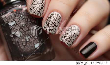 Close-up of a hand with a crackle-patterned neutral manicure and a dark, glittery accent nail, holding a matching bottle of polish. 138298363