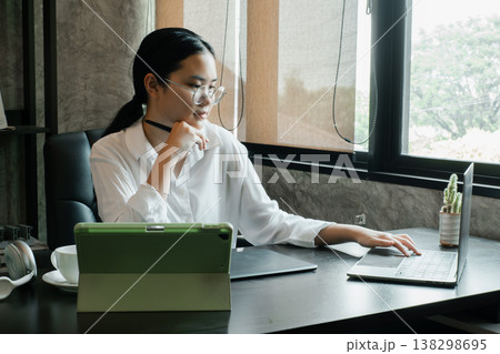 A young woman in a modern office setting, working on a laptop and tablet, surrounded by natural light from a large window. A young woman in a modern office setting, working on a laptop and tablet, surrounded by natural light from a large window. 138298695